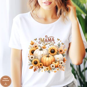 Peut inclure: T-shirt blanc avec un motif sur le thème de l'automne. Le motif comprend le mot "MAMA" dans une police décorative, entouré de citrouilles, de tournesols et de feuilles d'automne. Les citrouilles sont blanches et orange, et les tournesols sont jaunes.