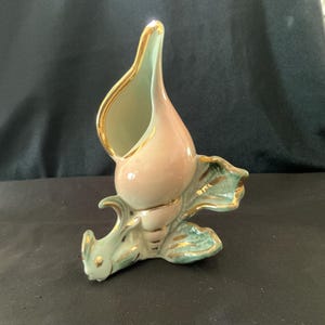 May include: A ceramic vase in the shape of a conch shell. The vase is primarily pink with a light green interior and accents. Gold trim outlines the edges and details. The base has a leaf-like design, also with gold trim.
