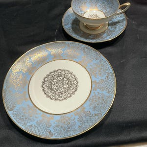 May include: A vintage tea set featuring a light blue and gold floral pattern. The set includes a teacup, saucer, and a larger plate. The teacup and saucer have gold trim, and the plate has a central floral design.