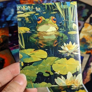 Puede incluir: Una impresión artística rectangular que representa una rana verde con ojos anaranjados en un estanque con nenúfares y flores blancas. La rana es el punto focal, rodeada de agua y follaje. Otras impresiones artísticas son visibles en el fondo.