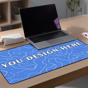 May include: A blue rectangular desk mat with white abstract designs and the text "YOU DESIGN HERE". A laptop, a pink mouse, a pink tumbler, and a white box are on the wooden desk.