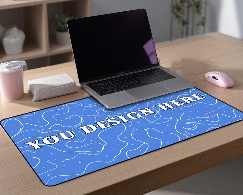 May include: A blue rectangular desk mat with white abstract designs and the text "YOU DESIGN HERE". A laptop, a pink mouse, a pink tumbler, and a white box are on the wooden desk.