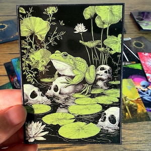 Puede incluir: Tarjeta rectangular negra con una ilustración gótica de una rana verde sobre una calavera en un estanque, rodeada de nenúfares y calaveras. La obra de arte utiliza tonos verde neón y blanco.