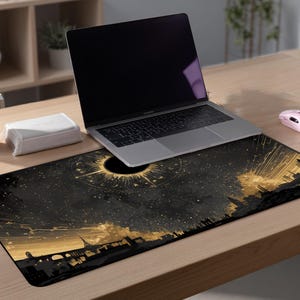 May include: A black and gold rectangular desk mat featuring a celestial design with a dark sun and a cityscape silhouette. A laptop, a pink mouse, a pink tumbler, and a white box are on a wooden desk.