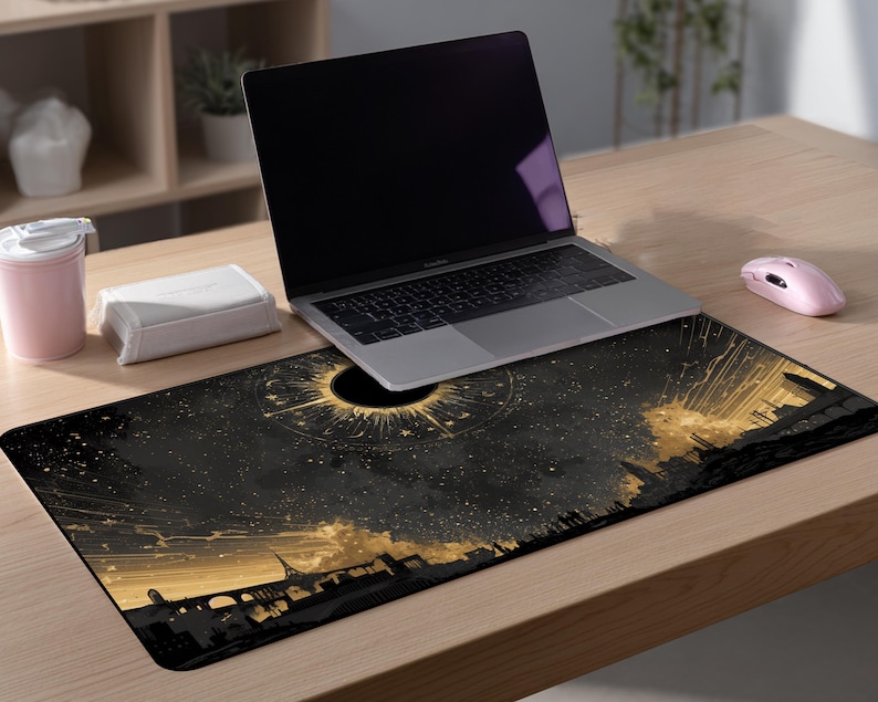 May include: A black and gold rectangular desk mat featuring a celestial design with a dark sun and a cityscape silhouette. A laptop, a pink mouse, a pink tumbler, and a white box are on a wooden desk.