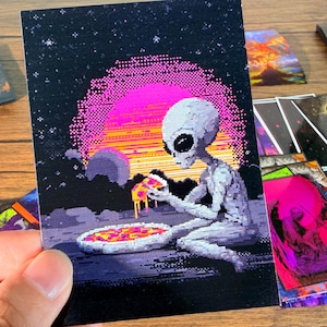 May include: A pixelated art print featuring an alien enjoying pizza against a backdrop of a purple sunset and starry sky. The alien is gray, and the pizza has red and yellow toppings. Other cards with similar designs are visible.
