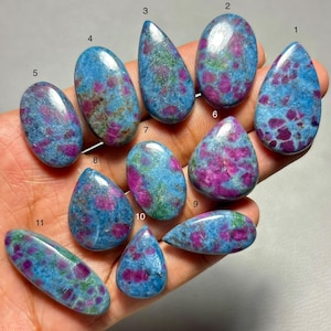 May include: A collection of eleven polished cabochon gemstones, each featuring a vibrant blue matrix speckled with distinct magenta and pink inclusions. The stones are numbered one through eleven and display various shapes, including teardrops and ovals.