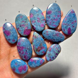 May include: Eleven polished cabochon gemstones featuring a blue matrix with embedded magenta and pink inclusions. Each stone is numbered from 1 to 11, displaying various oval and teardrop shapes with smooth, reflective surfaces and distinct color patterns.