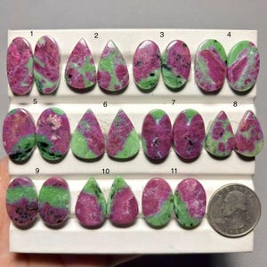 May include: A collection of eleven pairs of polished zoisite and ruby cabochons in oval and teardrop shapes, featuring vibrant magenta and green patterns. A United States quarter coin is included for scale on a white display stand with numbered rows.