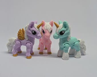 Könnte beinhalten: Drei Spielzeug-Einhörner in Pastellfarben: Lavendel, Rosa und Türkis. Jedes hat eine weiße Mähne und einen weißen Schweif, ein goldenes oder gelbes Horn und bewegliche Gelenke. Das Lavendel-Einhorn hat goldene Flügel.