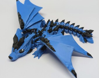 Könnte beinhalten: Eine blau-schwarze, 3D-gedruckte Drachenfigur. Der Drache hat große, bewegliche Flügel, einen langen Schwanz und schwarze Akzente entlang seines Rückens und seiner Flügel. Die Figur liegt auf einer weißen Oberfläche.