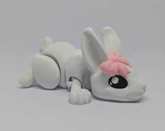 Könnte beinhalten: Eine weiße, bewegliche Hasenfigur mit einer rosa Blume auf dem Kopf. Der Hase hat schwarze Augen und liegt. Die Figur besteht aus einem glatten, matten Material.