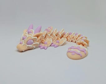 Könnte beinhalten: Detaillierte, bewegliche Drachenfigur in Pfirsich-, Lavendel- und Weißtönen. Der Drache hat einen detaillierten Kopf, spitze Hörner und einen segmentierten Körper. Ein kleines, rundes, dekoriertes Stück befindet sich neben dem Drachen. Die Figur steht auf einer weißen Oberfläche.