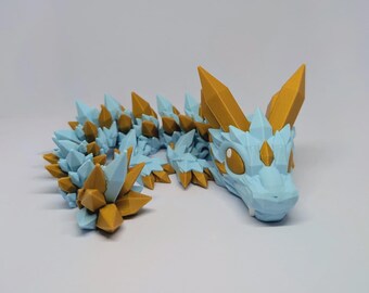 Könnte beinhalten: Eine bewegliche Drachenfigur in Blau und Gold. Der Drache hat einen hellblauen Körper mit goldenen Akzenten und kristallartigen Stacheln entlang des Rückens. Der Kopf hat große, runde Augen und spitze Hörner. Die Figur ist aus Kunststoff.