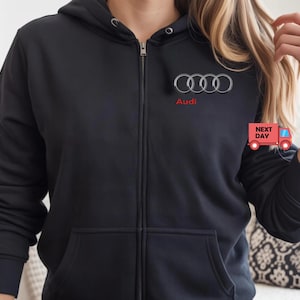 May include: Black zip-up hoodie with the Audi logo and text in red. The hoodie has a hood with drawstrings, a full-length zipper, and two front pockets. The Audi logo is a set of four interlocking rings.
