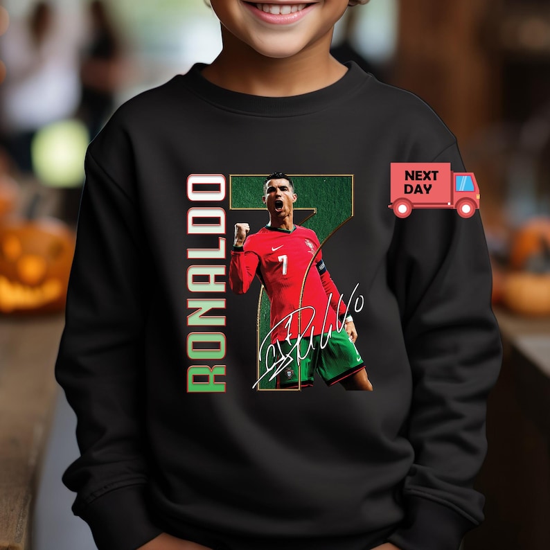 May include: Black sweatshirt featuring a graphic of Cristiano Ronaldo in a red jersey with the number 7. The word "RONALDO" is printed vertically in green and red. Includes a signature.