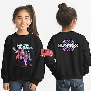 May include: Black sweatshirt featuring the text "KPOP DEMON HUNTERS" with a graphic of three figures. The back of the sweatshirt displays the word "HUNTRIX" with a colorful, intertwined design. The sweatshirt is worn by a child.