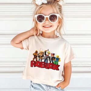 May include: A beige t-shirt featuring a colorful graphic of Roblox characters and the word "ROBLOX" in red block letters. The shirt is worn by a young child wearing white sunglasses.