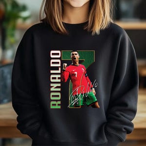 May include: Black sweatshirt featuring a graphic of a soccer player in a red jersey with the number 7. The image includes the player's signature and the name "RONALDO" in green and red.