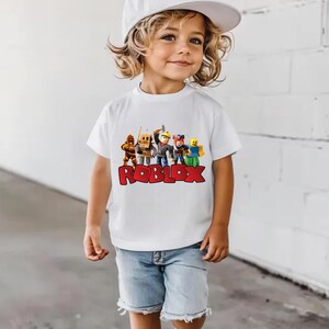 May include: A white t-shirt featuring a colorful graphic of Roblox characters and the Roblox logo in red. The shirt is worn by a young child, paired with denim shorts and a white baseball cap.
