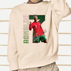 May include: Cream-colored sweatshirt featuring a graphic of a soccer player in a red jersey and green shorts. The player is positioned in front of a large green number 7. The word "RONALDO" is printed vertically in red and green. The player's signature is also visible.
