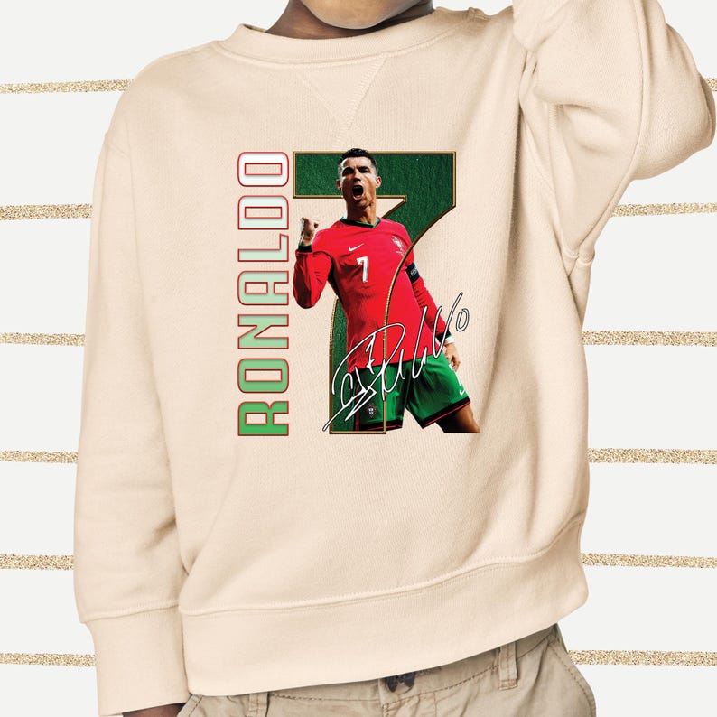 May include: Cream-colored sweatshirt featuring a graphic of a soccer player in a red jersey and green shorts. The player is positioned in front of a large green number 7. The word "RONALDO" is printed vertically in red and green. The player's signature is also visible.
