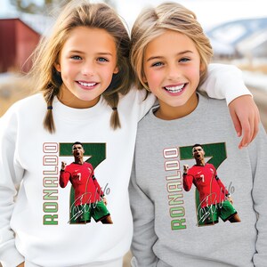 May include: Two children wearing white and gray sweatshirts. Each sweatshirt features a graphic of a soccer player in a red jersey with the number 7, green shorts, and the name "RONALDO" in green and red.