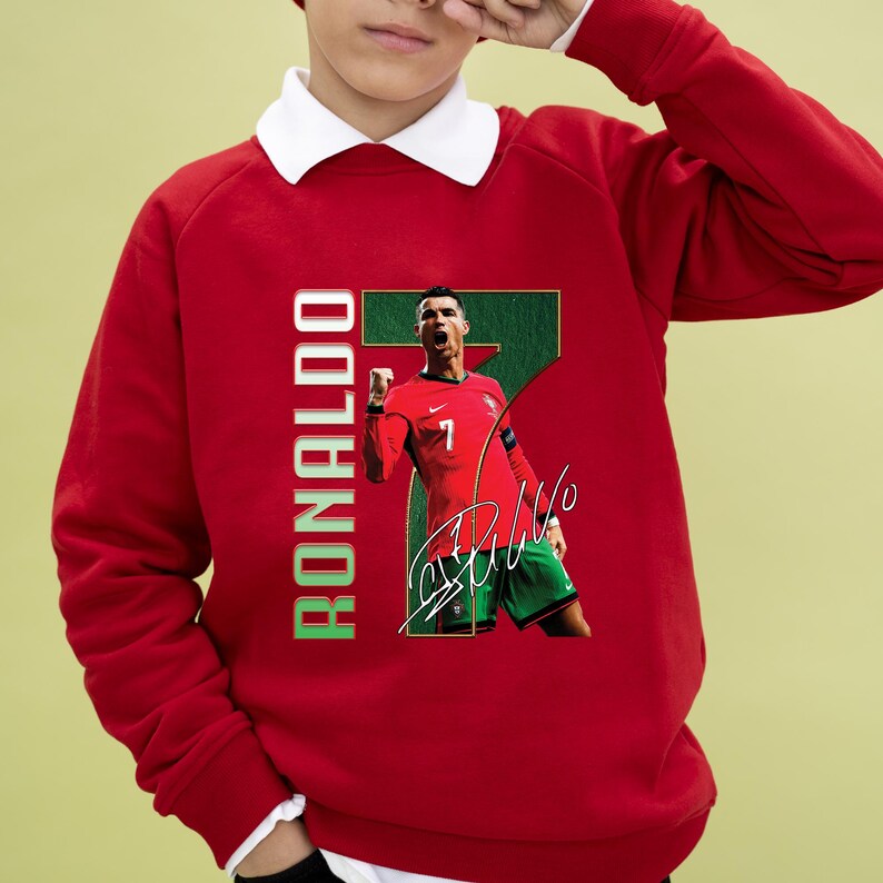 May include: Red sweatshirt featuring a graphic of a soccer player in a red jersey with the number 7, green shorts, and a signature. The word "RONALDO" is printed vertically in green and white. The sweatshirt has a white collar.