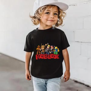 May include: A black t-shirt featuring a colorful graphic of Roblox characters and the word "ROBLOX" in red. The shirt is worn by a child wearing a white baseball cap and blue jeans.