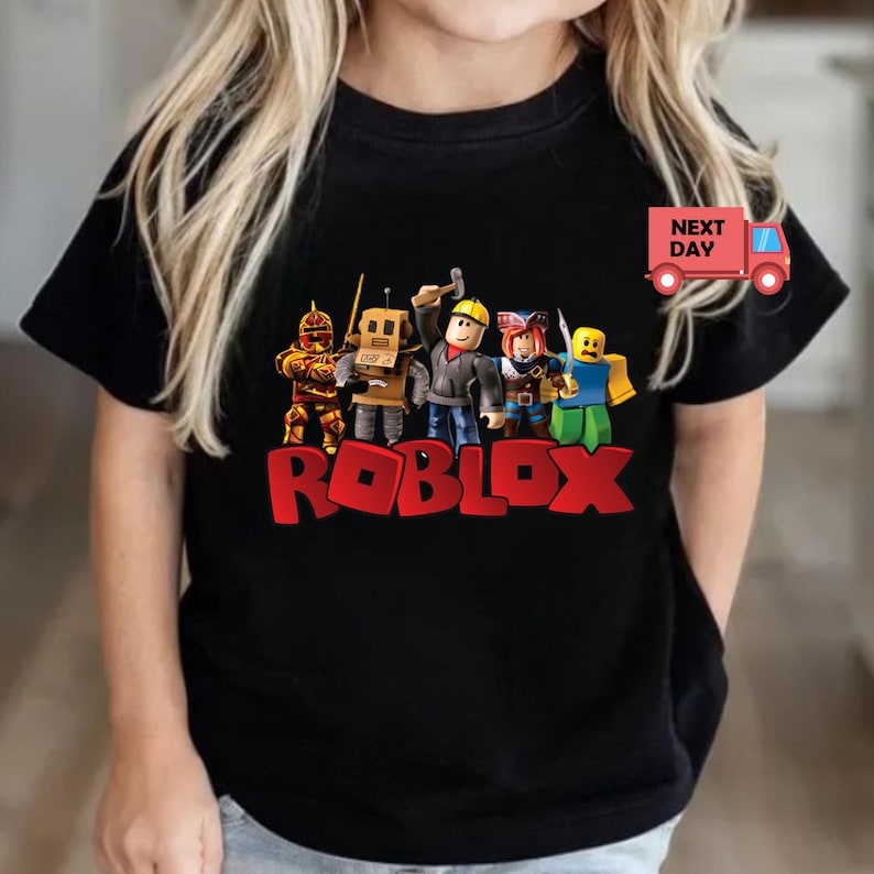 May include: Black t-shirt featuring the word "ROBLOX" in red block letters with a group of cartoon characters above it. The characters are in various outfits and poses. A red truck with the words "NEXT DAY" is on the right.