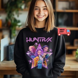 May include: Black sweatshirt featuring the word "HUNTRAX" in purple, with a graphic of three women and a blue cat inside a star. The sweatshirt has a crew neck and long sleeves. A red truck with the words "NEXT DAY" is in the upper right corner.