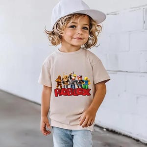 May include: A beige t-shirt featuring a colorful graphic of Roblox characters and the Roblox logo in red. The shirt is worn by a young child, paired with blue jeans and a white baseball cap.