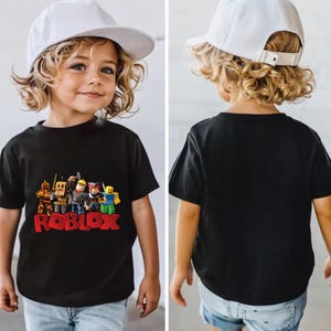 May include: Black short-sleeved t-shirt featuring a colorful graphic of Roblox characters and the Roblox logo in red. The child is wearing a white baseball cap. The back of the shirt is plain black.