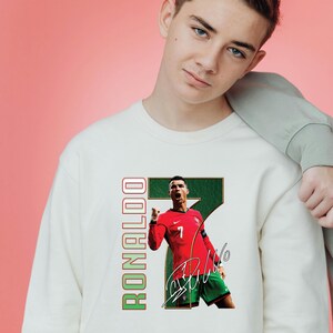 May include: A white sweatshirt featuring a graphic of a soccer player in a red jersey with the number 7. The word "RONALDO" is printed vertically in green and red. The player is celebrating a goal.