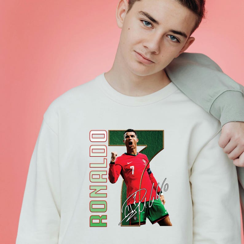 May include: A white sweatshirt featuring a graphic of a soccer player in a red jersey with the number 7. The word "RONALDO" is printed vertically in green and red. The player is celebrating a goal.