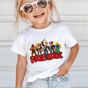 May include: White t-shirt featuring a colorful graphic of Roblox characters and the Roblox logo in red. The shirt is short-sleeved and appears to be made of cotton. The design includes several animated figures.
