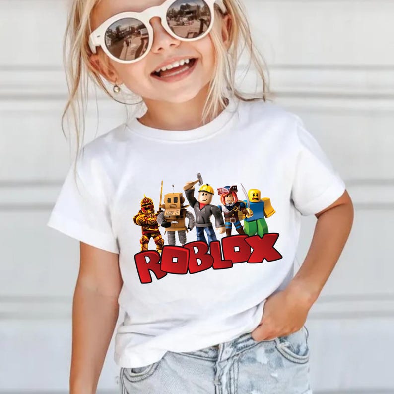 May include: White t-shirt featuring a colorful graphic of Roblox characters and the Roblox logo in red. The shirt is short-sleeved and appears to be made of cotton. The design includes several animated figures.