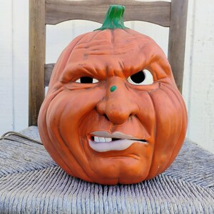 Könnte beinhalten: Orange Halloween-Kürbisdekoration mit einem grimmigen Gesicht. Der Kürbis hat einen grünen Stiel und ein detailliertes, ausdrucksstarkes Gesicht mit gerunzelter Stirn, einem schiefen Mund und einem kleinen grünen Punkt auf der Nase. Der Kürbis sitzt auf einem Stuhl.