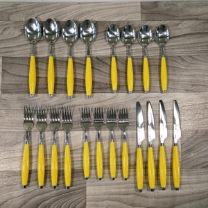 May include: A flat lay of a vintage silverware set with yellow handles. The set includes spoons, forks, and knives. The spoons and forks are arranged in two rows, with the knives in a separate row. The silverware is made of stainless steel.