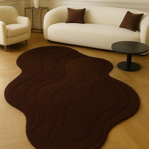 May include: A dark brown, organically shaped rug with a wavy design. The rug is placed on a light-colored wooden floor, and is surrounded by neutral-toned furniture, including a sofa, armchair, and side table. The rug's dimensions are approximately 72 inches by 50 inches.