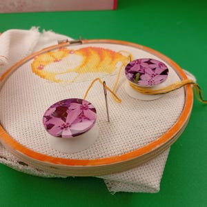 May include: A cross-stitch project in progress, featuring an orange and yellow cat design on white fabric stretched within an orange embroidery hoop. Two pink floral-patterned needle minders and a needle with yellow thread are also visible.