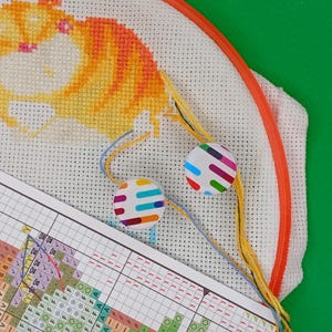 May include: A cross-stitch project in progress, featuring a cartoon cat design in orange and yellow. The fabric is stretched in an orange hoop, with colorful threads and two round, patterned magnets. A chart with color-coded symbols is also visible.