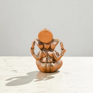May include: A bronze-colored statue of the Hindu deity Ganesha, seated in a meditative pose. The sculpture features intricate details, including the elephant head, multiple arms, and a round headdress. The statue is on a white surface.