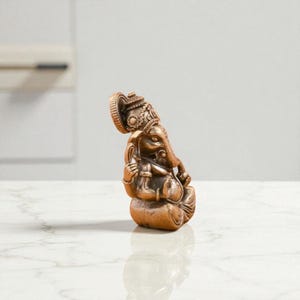 May include: A bronze-colored statue of the Hindu deity Ganesha, seated in a meditative pose. The statue features intricate details, including a large head with elephant features, ornate headdress, and multiple arms. The statue is on a white surface.