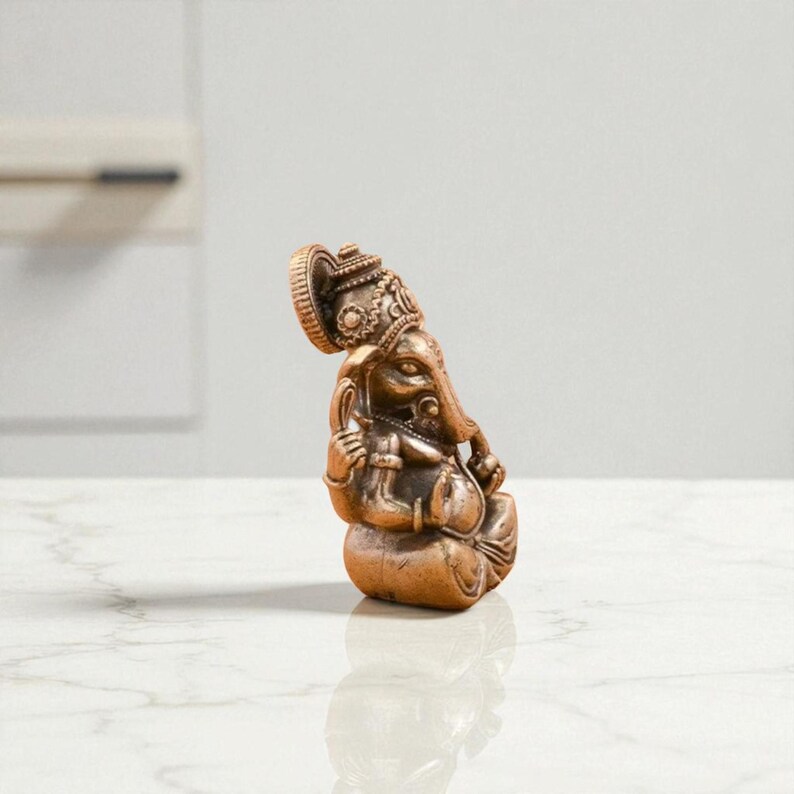 May include: A bronze-colored statue of the Hindu deity Ganesha, seated in a meditative pose. The statue features intricate details, including a large head with elephant features, ornate headdress, and multiple arms. The statue is on a white surface.