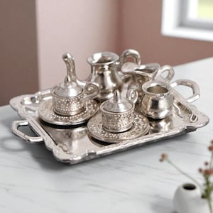 Puede incluir: Un juego de té plateado en miniatura sobre una bandeja a juego. El juego incluye una tetera, una crema, un azucarero y dos tazas de té con platillos. La bandeja tiene asas y un borde decorativo. El juego es de metal brillante.