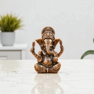May include: A bronze-colored statue of the Hindu deity Ganesha, seated in a meditative pose. The statue features intricate details, including a large elephant head, multiple arms holding various objects, and ornate headdress. The statue is on a white surface.