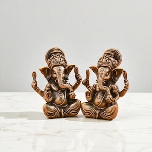 May include: Two bronze-colored Ganesh statues. Each statue depicts the Hindu deity Ganesh in a seated position, holding a small object in each hand. The statues have detailed ornamentation and are set against a neutral background.