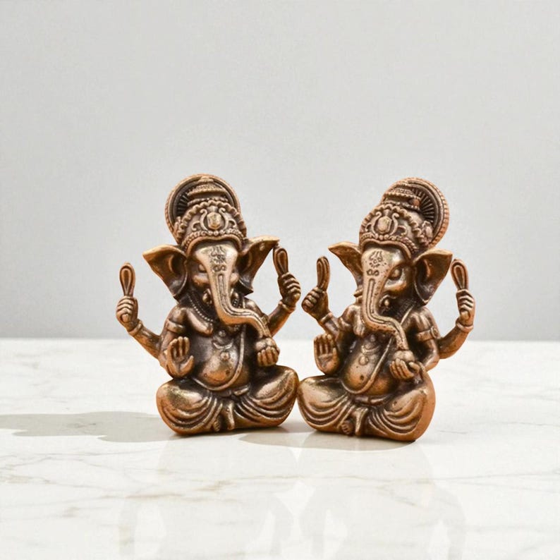 May include: Two bronze-colored Ganesh statues. Each statue depicts the Hindu deity Ganesh in a seated position, holding a small object in each hand. The statues have detailed ornamentation and are set against a neutral background.
