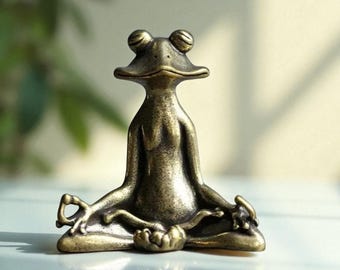 May include: A bronze-colored frog figurine in a seated yoga pose. The frog has large, round eyes and is positioned with its legs crossed and arms outstretched. The figurine is made of metal and has a detailed, textured surface.